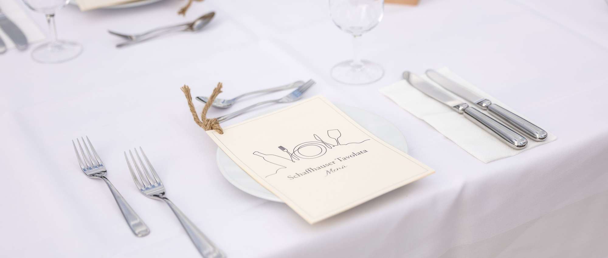 Auf einem weiss gedeckten Tisch liegt auf dem Teller ein Menü-Heftchen mit der Aufschrift Schaffhauser Tavolata und einer Linienzeichnung.
